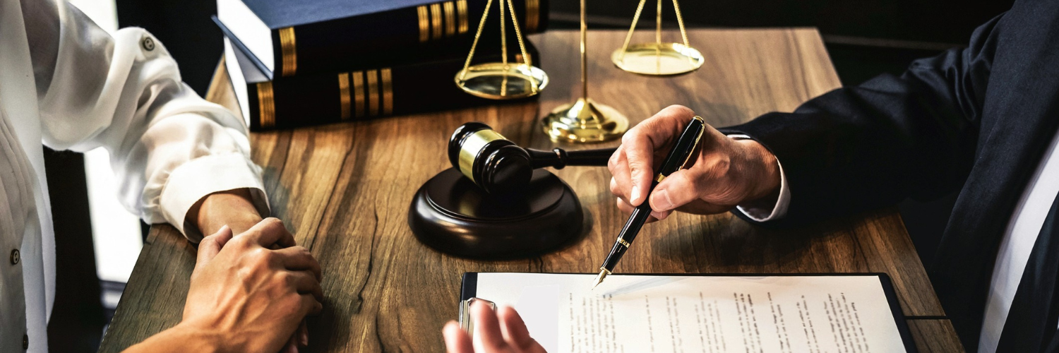 Photo of two people at a desk, with a document between them and a judge's gavel and scales on the desk